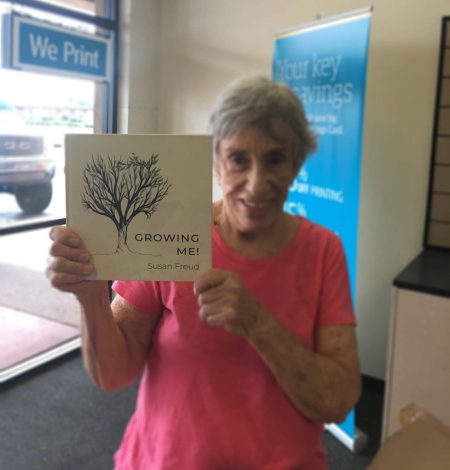 A woman smiling and holding up a copy of the book Growing Me! by Susan Freud inside a print shop, with store signage visible in the background.