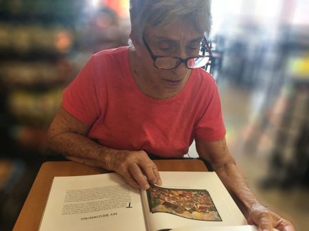 A woman wearing glasses and a pink shirt sits at a table reading an open book, focusing on a page titled “My Beginning” with an illustration on the right side. The background is softly blurred.